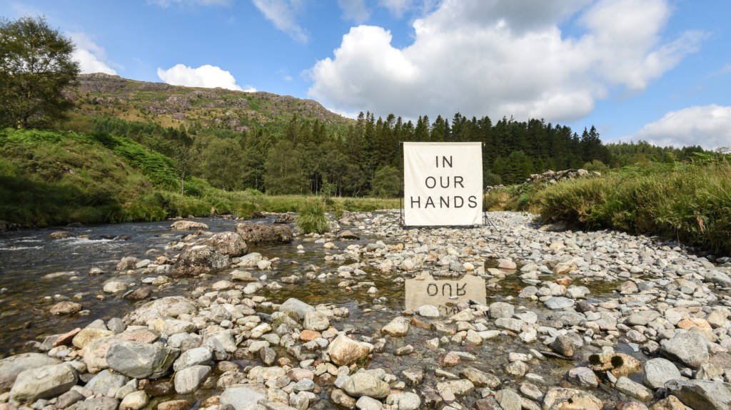 A large white canvas erected on a river bed in the Duddon Valley. The canvas has the words IN OUR HANDS written on it in black.