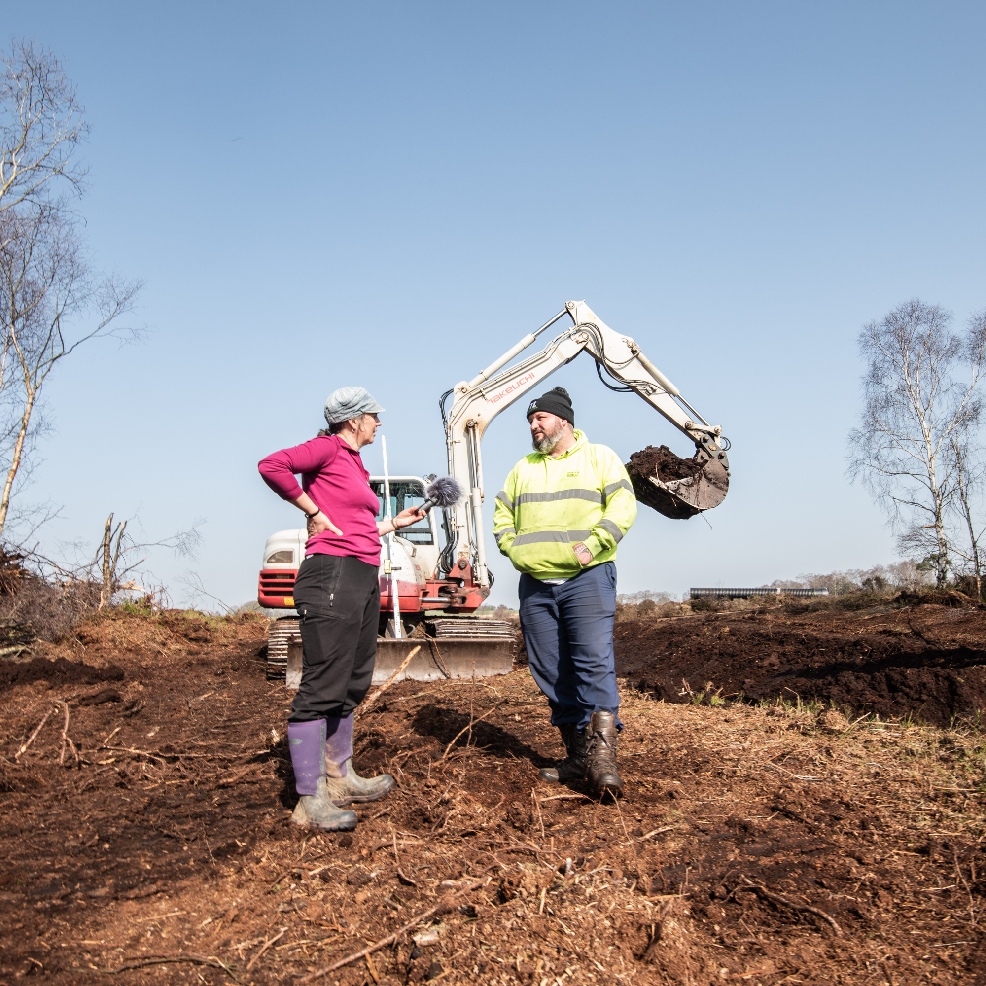 A woman wearing a bright pink top is holding a microphone and is talking to a man wearing a high vis jacket and a beany hat. It is a bright sunny day. In the background there is a large digger with its bucket raised in the air.