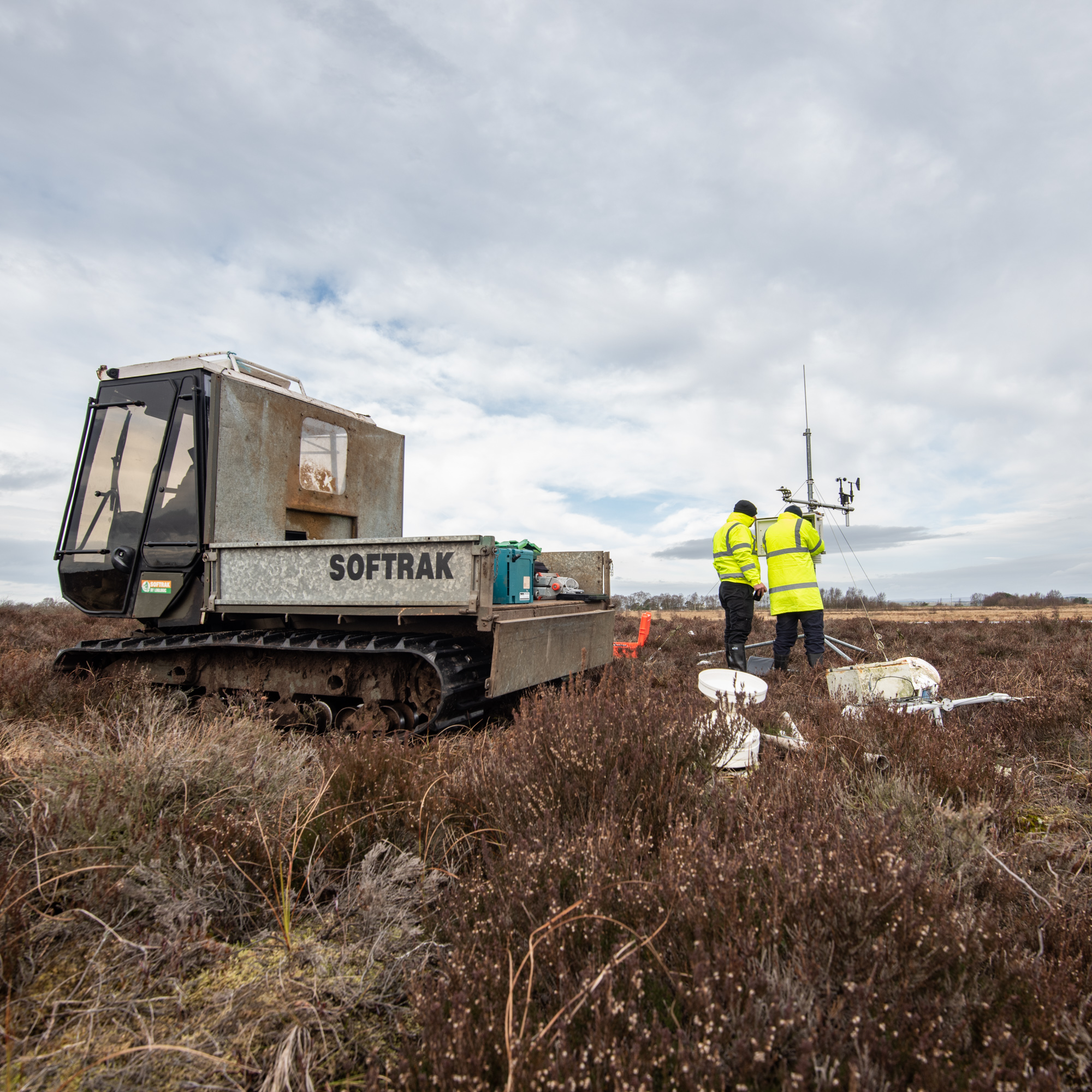 Two men looking at a small weather station on the bog. In the foreground there is a caterpillar tracked vehicle.