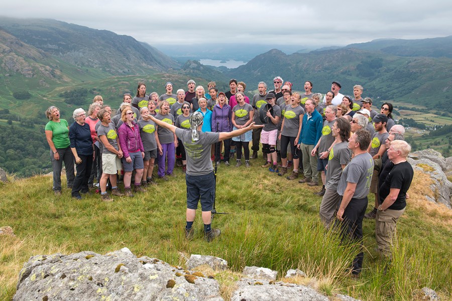 A choir sings in the open air: they are standing on Thorneythwaite Fell, with lakes and mountains behind them