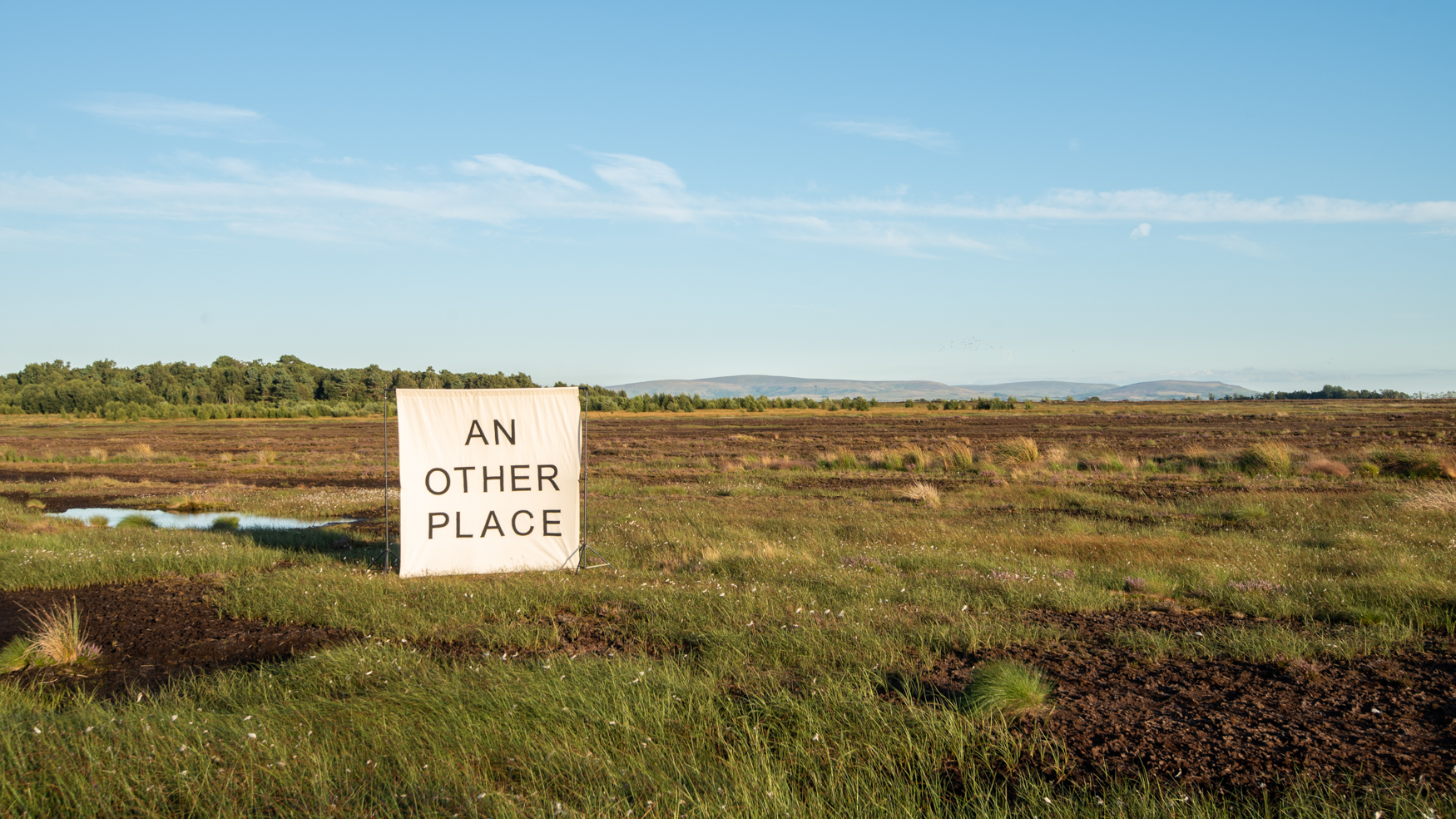 A large canvas set on the flat ground of a peat bog. The words on the canvas read 'AN OTHER PLACE'