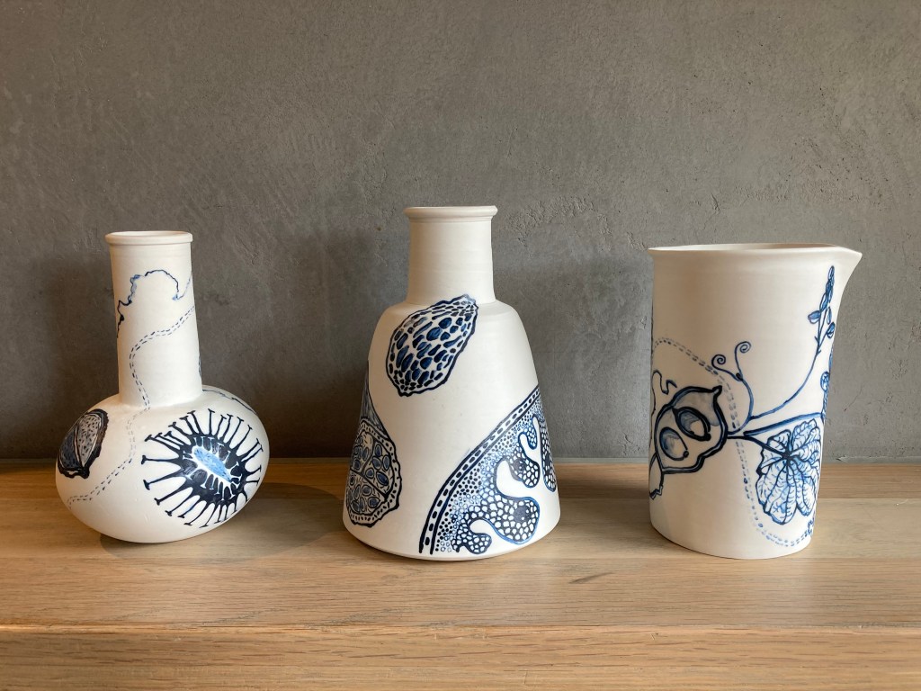 Three ceramic pots which are white with blue drawings on them representing food