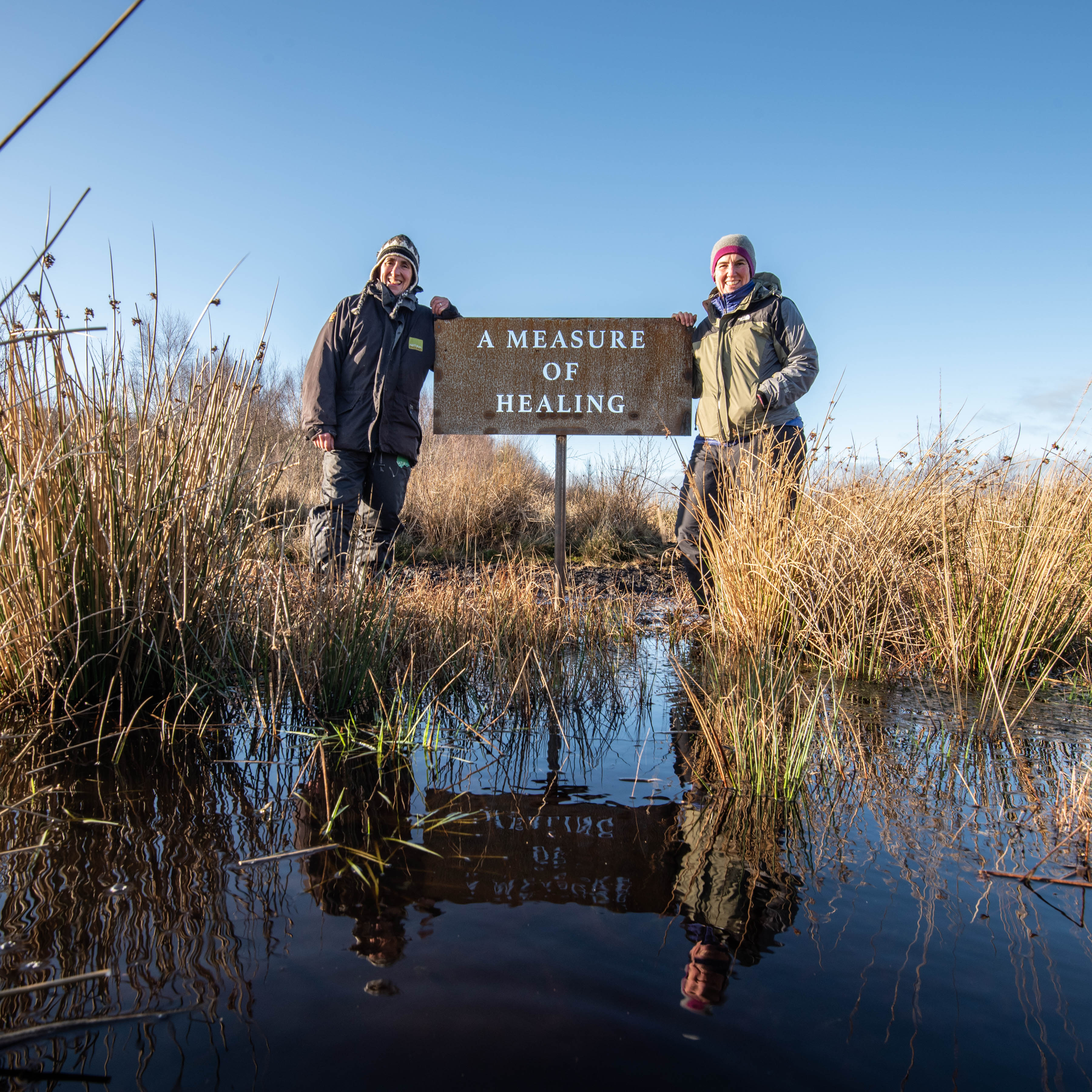 Two women stand either side of a steel sign that has words cut into it reading 'A Measure of Healing'