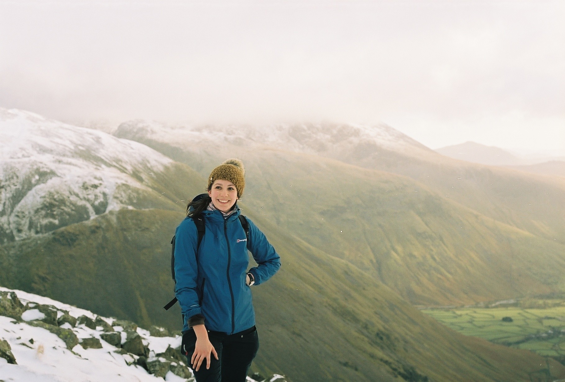 A woman stands on a hilltop, with snowy hills behind. She is wearing a yellow hat and a blue jacket.
