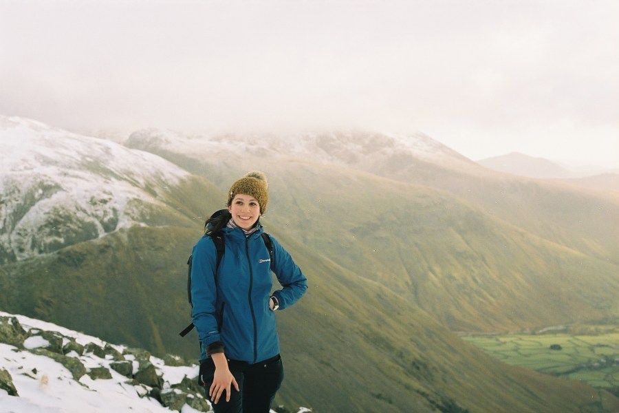 A woman stands on a hilltop, with snowy hills behind. She is wearing a yellow hat and a blue jacket.