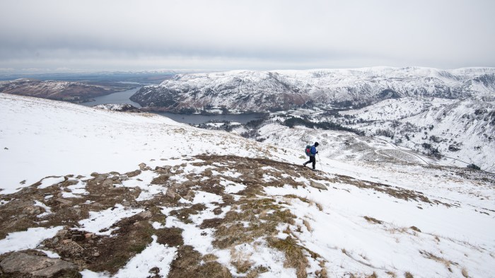 A snowy scene of hills with a lake in the distance and a single walker heading down the path in the centre of the frame