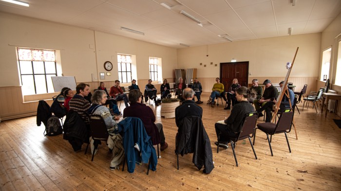 Twenty people are sitting in a wide circle, in a village hall, facing inwards.