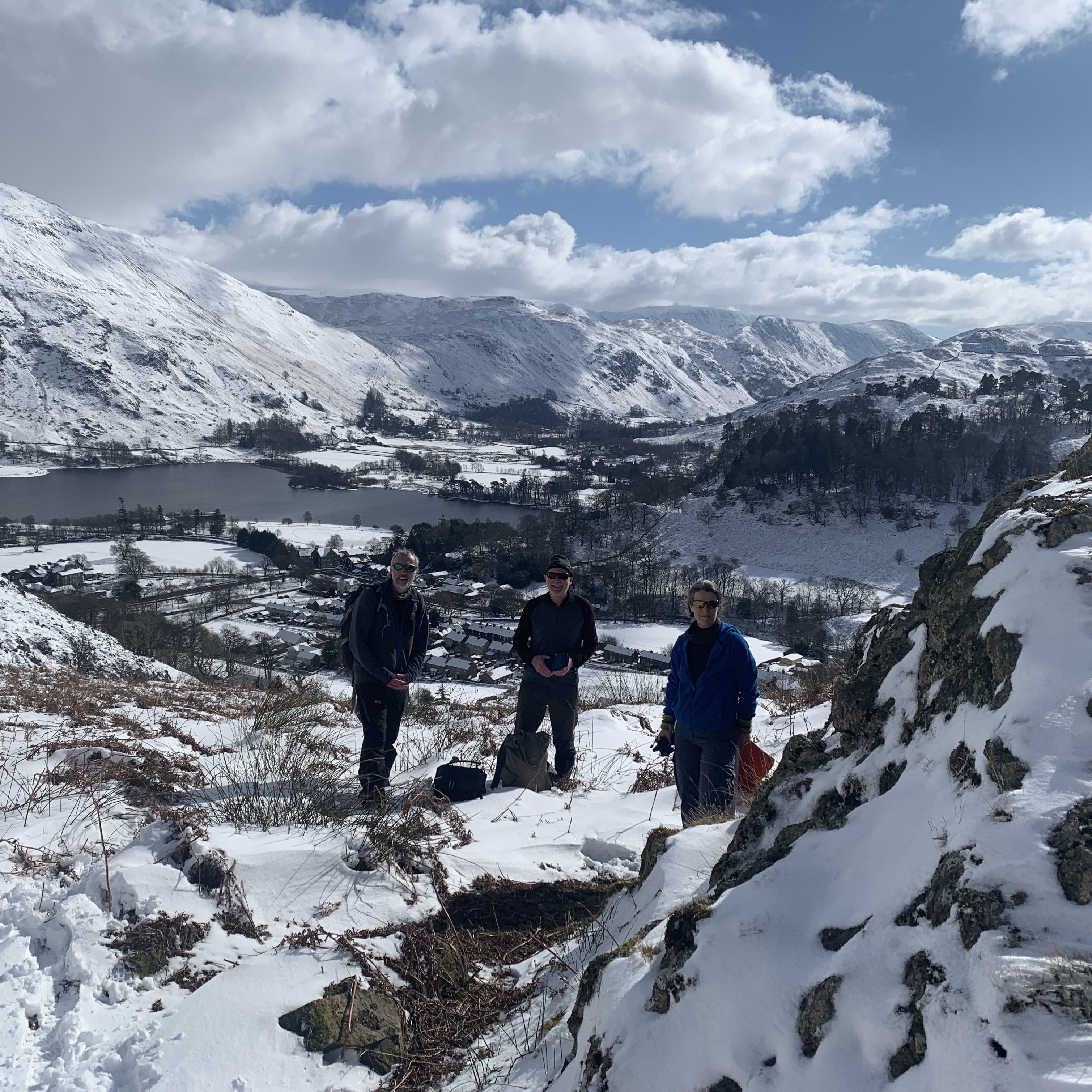 Three people stand on a hill above a lake on a sunny day: the land is covered by snow