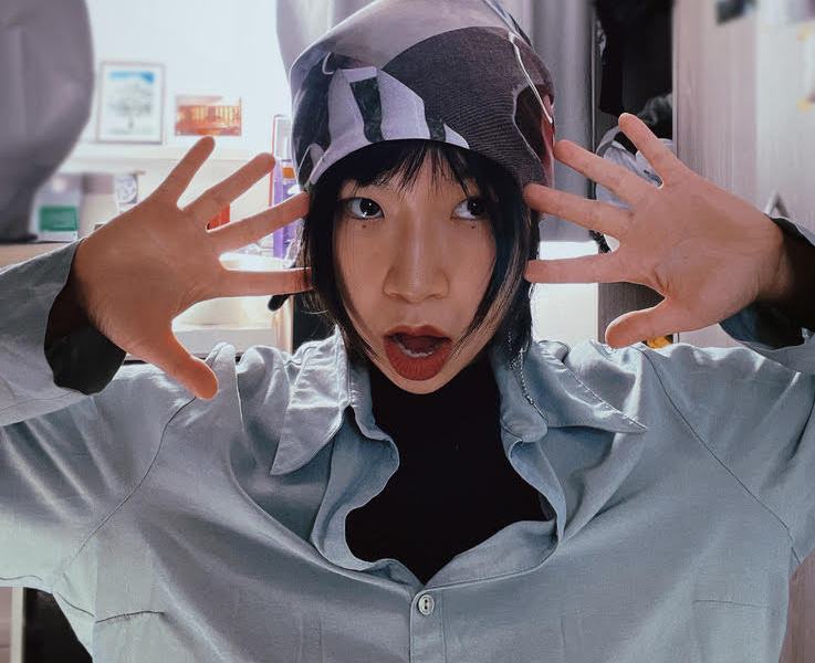 A woman with a headscarf faces the camera with hands either side of her head, fingers splayed.