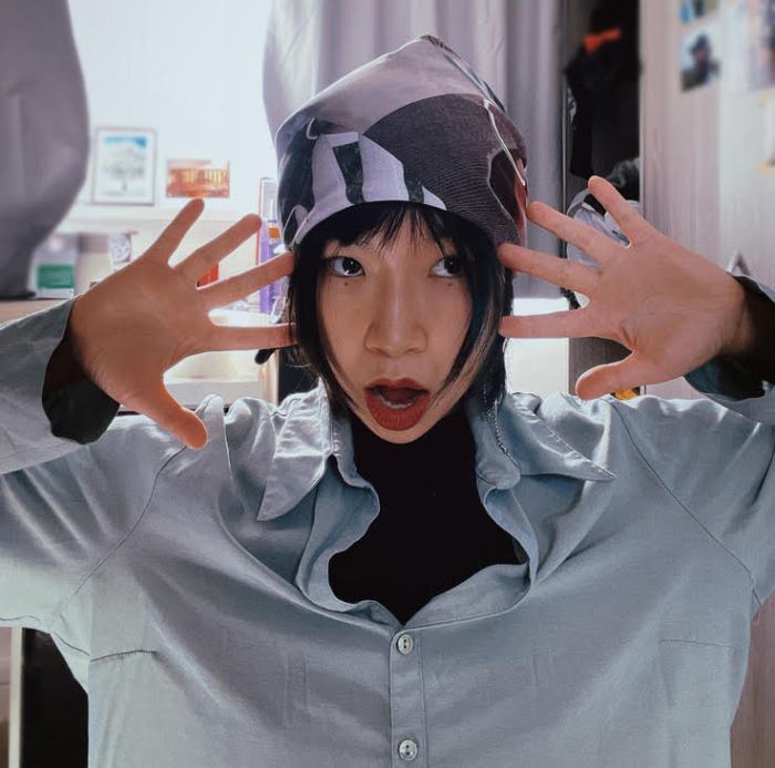 A woman with a headscarf faces the camera with hands either side of her head, fingers splayed.