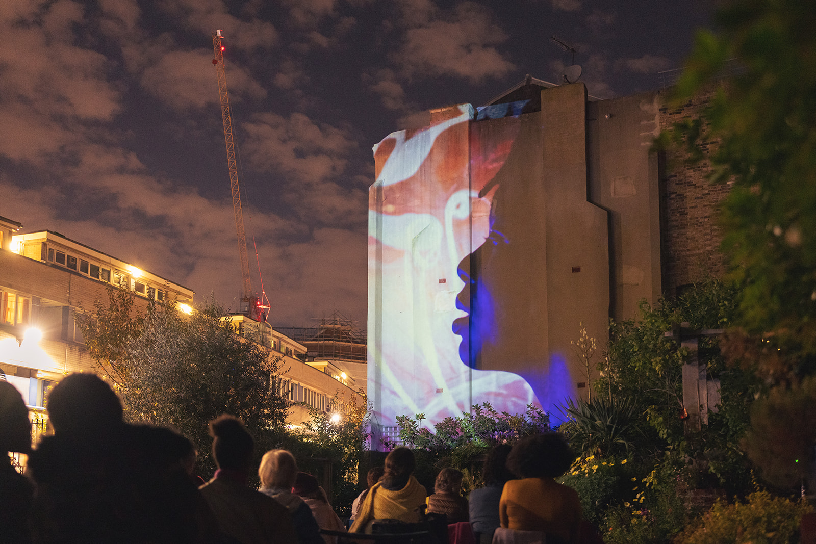 A silhouette of a face is visible on the side of a building, as a projection, with coloured background. A crowd of people watch.