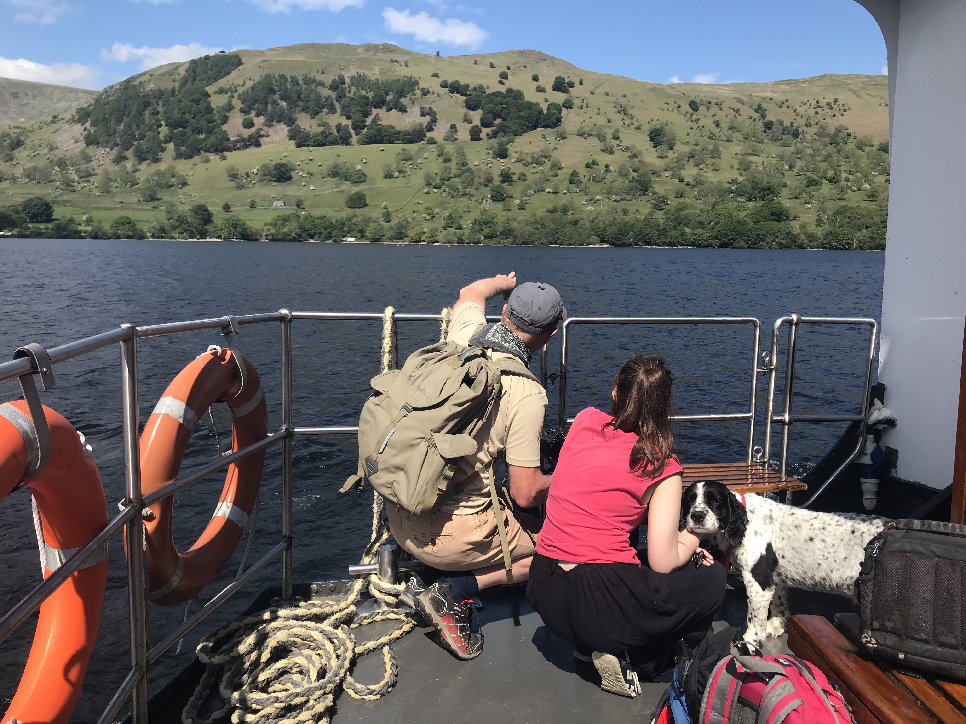 Two people look over a lake from the deck of a boat. A black and white dog looks at the camera. There are hills in the background
