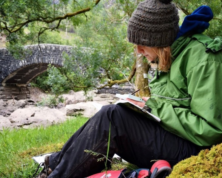 A woman in a bobble hat sits on a river bank, sketching