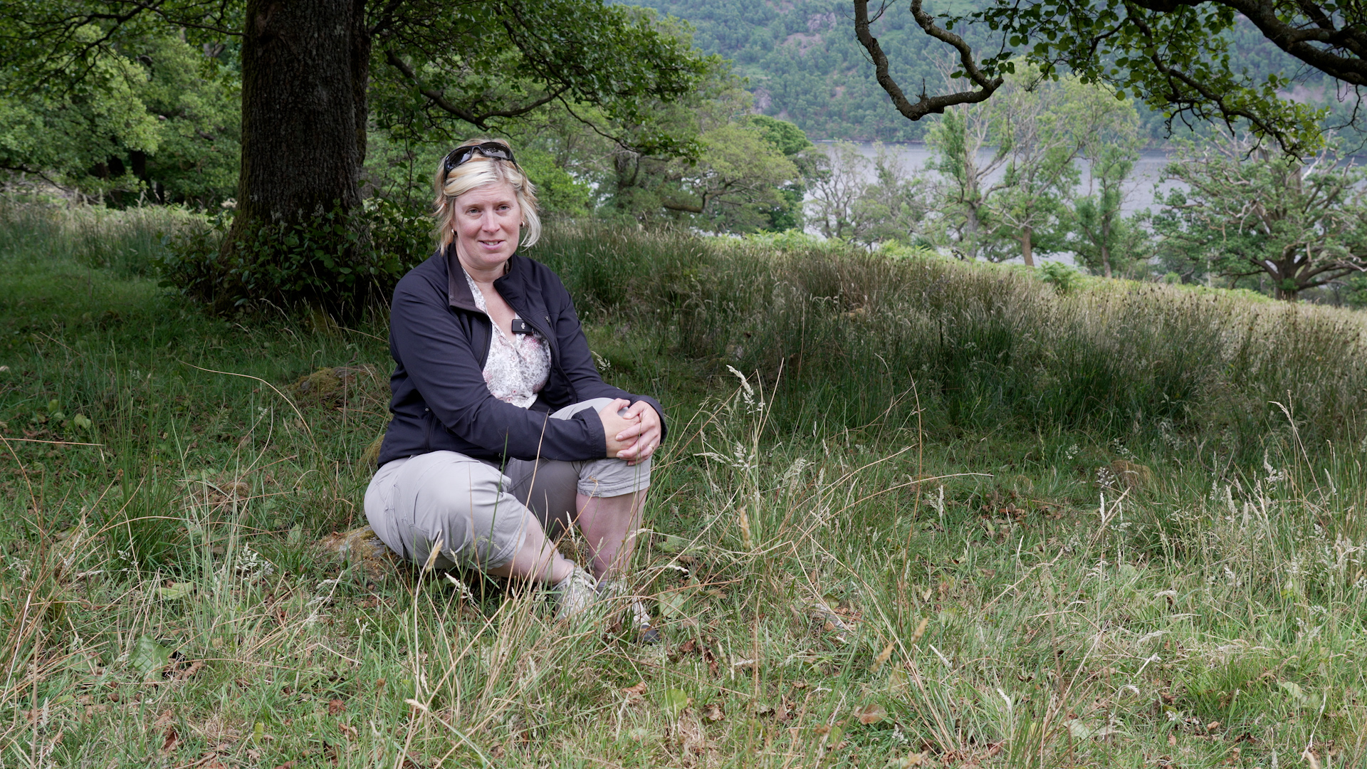A woman sits in the shade of a tree, with a lake visible in the background