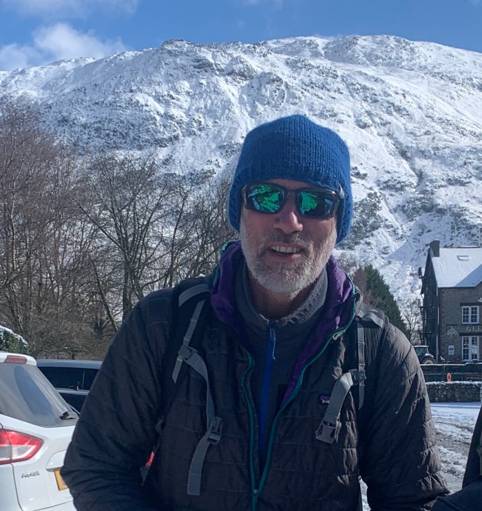 A man in a blue hat, and wearing reflective sunglasses, stands in sunshine, with a snow-covered mountain behind him