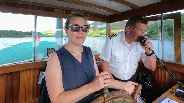 A woman holds the wheel of a boat, while a skipper in uniform speaks into the radio