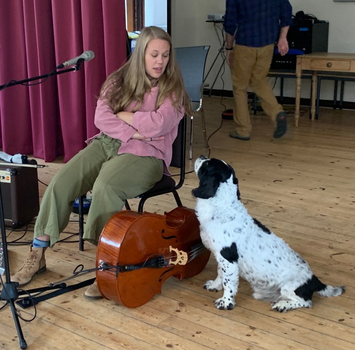 A woman sits on a chair, talking to a black and white dog, with a cello resting between them.