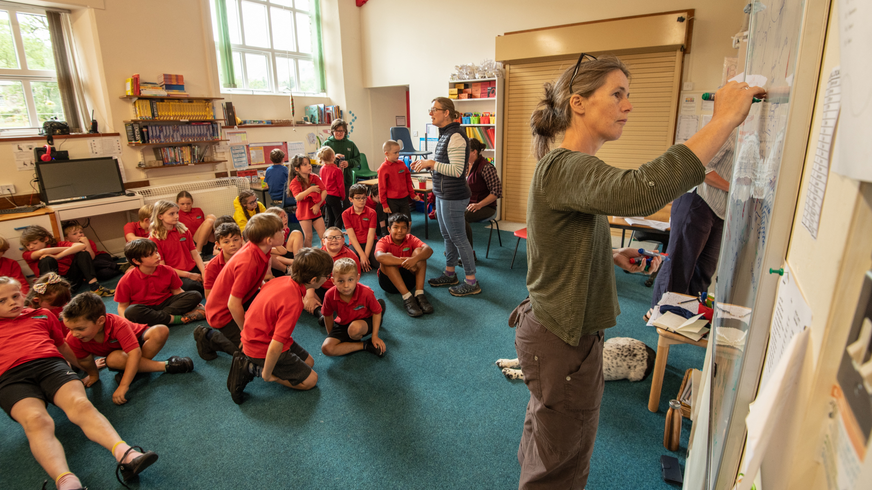 A woman marks a whiteboard in a classroom; children sit on the floor