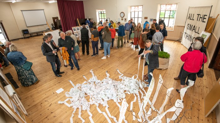 A group of people gather around artworks, with a woman in the foreground looking through long threads of paper