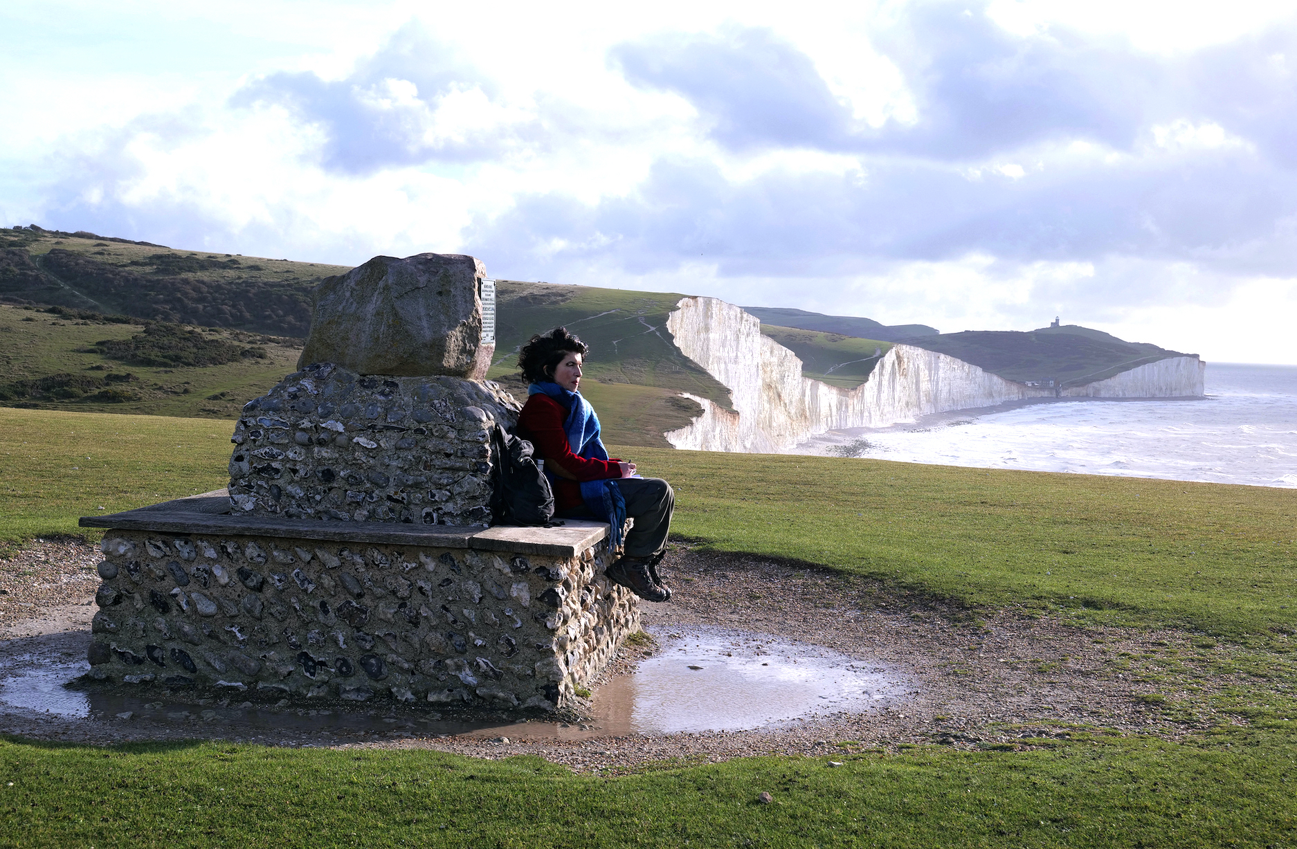 A woman sits on a small stone monument. In the background are white cliffs and the sea.