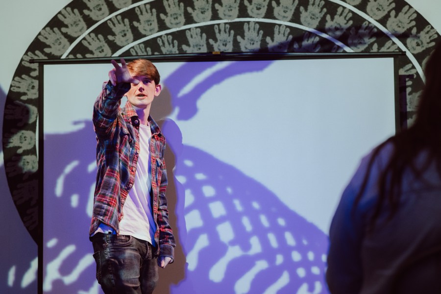A young man stands in front of an image on a screen, and gestures towards the camera