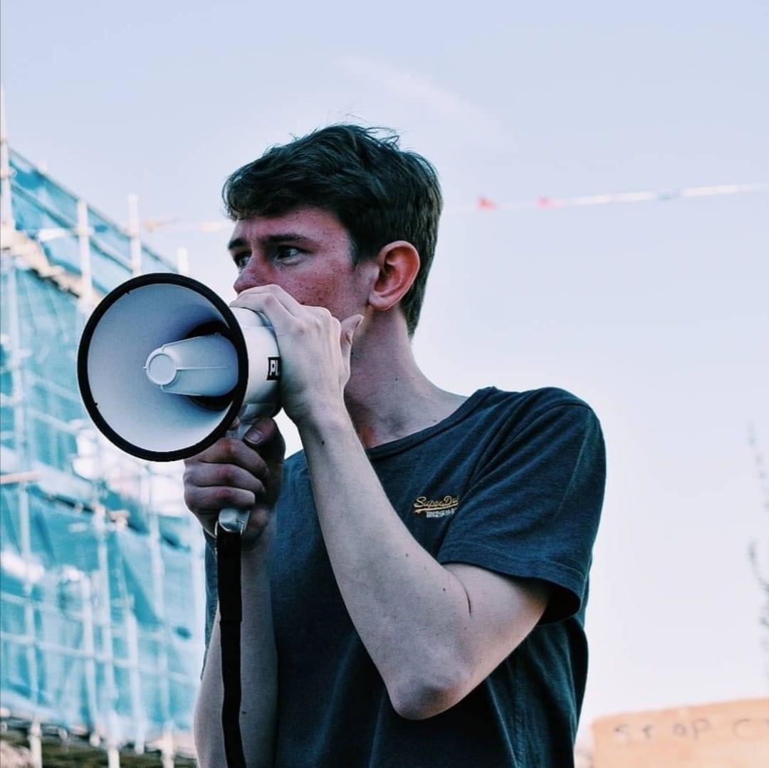 A young man holding a megaphone to his mouth, with a building behind.
