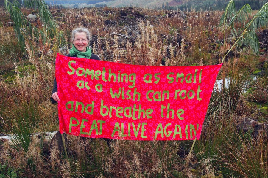 A woman stands holding a red banner with the words: 'Something as small as a wish can root and breathe the peat alive again'
