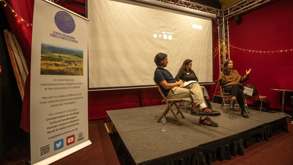 Three people sitting on a stage, one is talking and raising her hands.