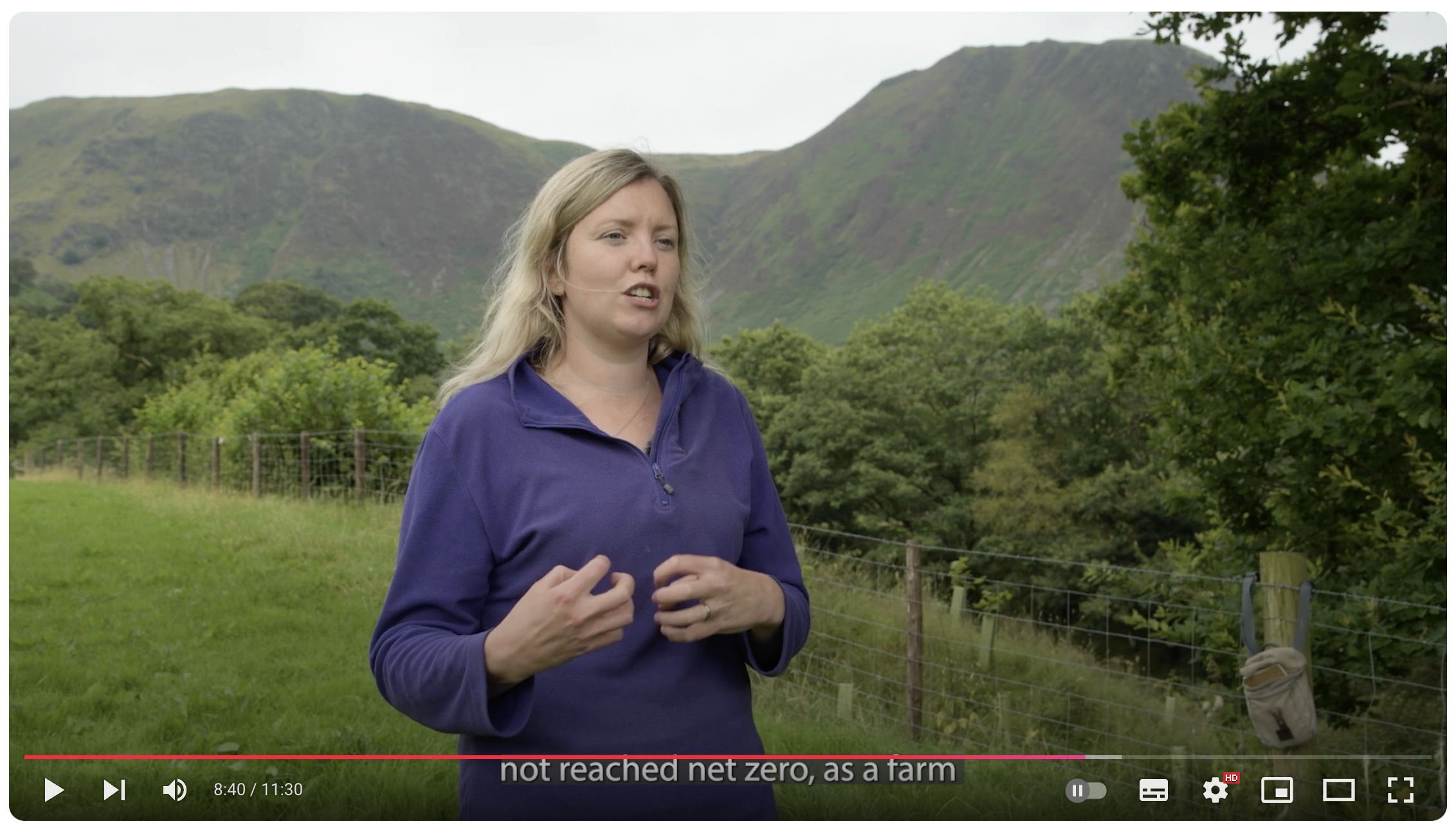 A woman speaks while standing in a field, with trees and a hill behind her