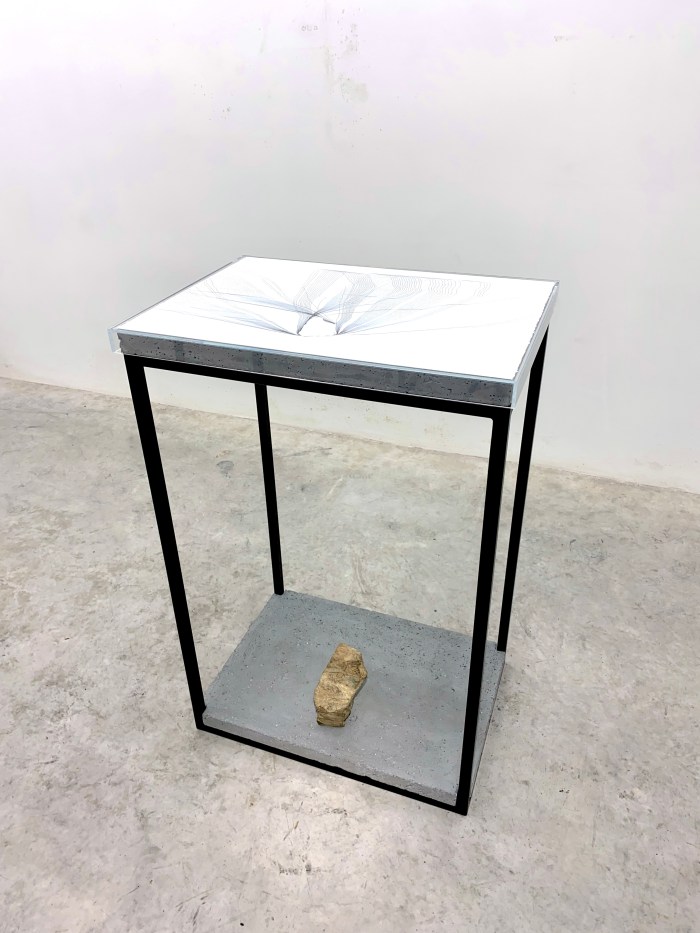 A table with a black frame and concrete slabs at the bottom and top. A rock is stood on the bottom concrete slab. There is a drawing on top of the table and a clear case over the top. The table stands in a grey room.
