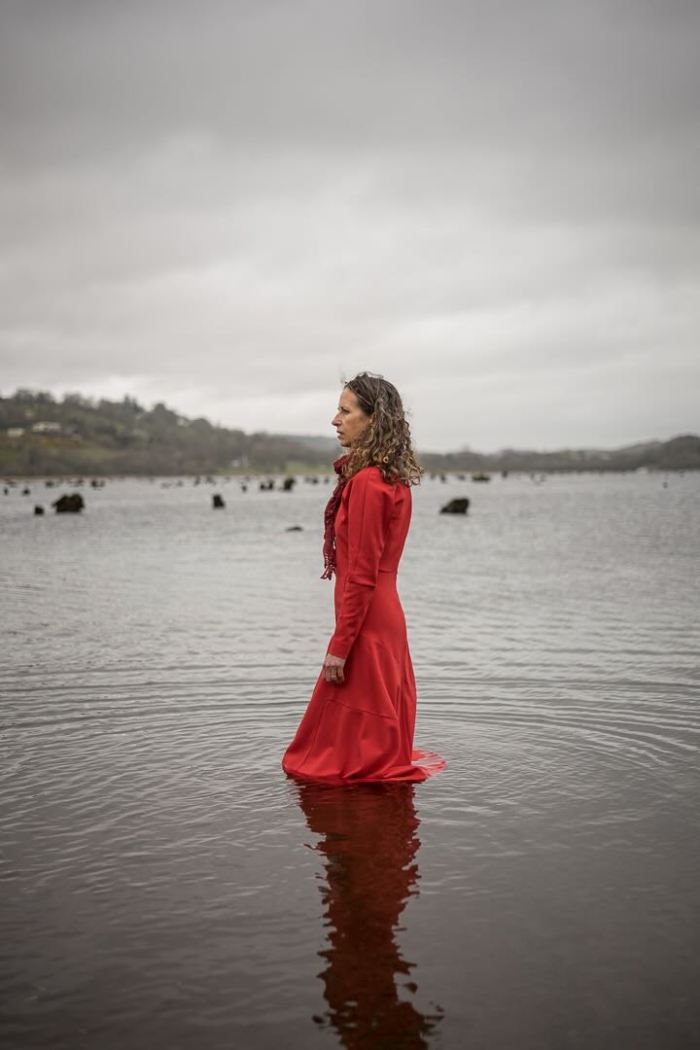 a woman in a red dress stands in a pool of water, with grey sky