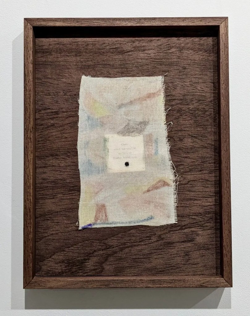 A dark wooden frame containing a small patch of fabric, there are faint blocks of colour on the fabric and there appears to be a white square underneath with words printed on it.