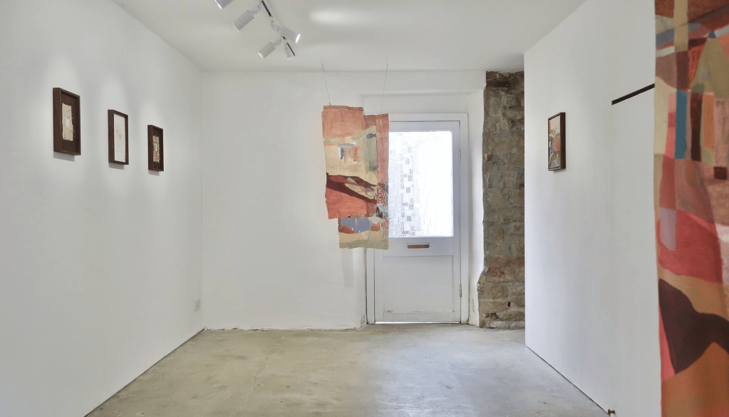 An exhibition space with white walls, a concrete floor, and a door in the back with a window. There is a corridor at the back of the room with grey stone walls. On the left wall there are three frames images. Hanging by the door is a piece made from remnant textiles with sandy pink tones. Another similar textile piece is hung on the right hand wall, so close to the front of the image you can only see a little bit of it.