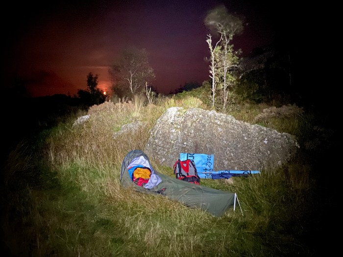 A camp at night time with a small 1-man tent, rucksack, and some outdoor gear in a rocky, grassy area with a few trees. There is a red glow in the sky and the silhouette of trees in the distance. The camp is lit up by a torch while the area around it remains dark.
