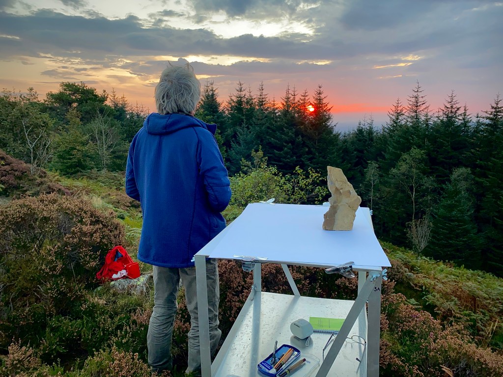 A man stands looking at the sunrise through some trees. There is a table beside him with a rock, and paper ready for sketching.