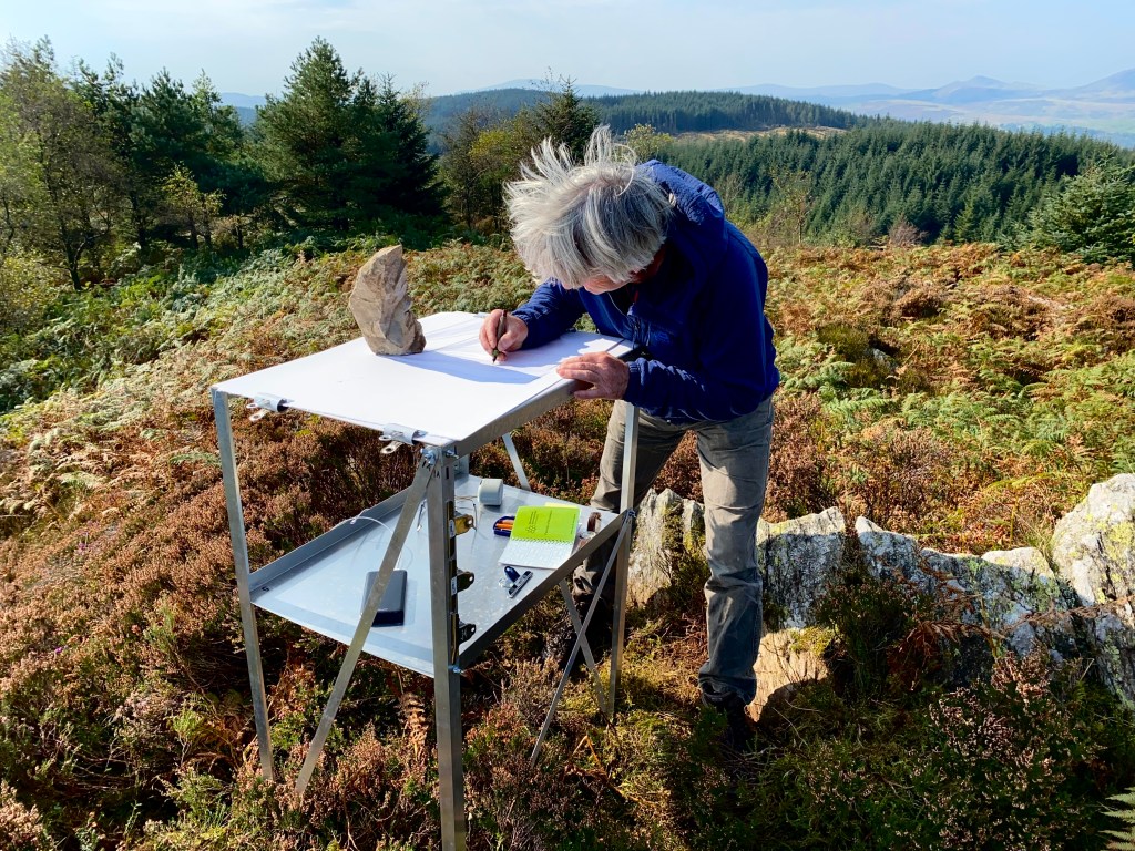 A man stands on a hill drawing at a metal table. On the table is a tall rock, he is drawing around its shadow. In the distance there are other hills, and in the mid ground there are lots of trees.