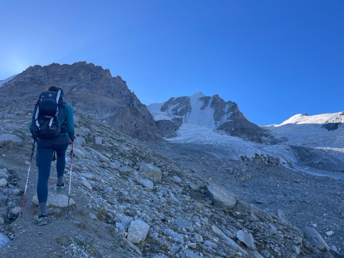 A woman walks across a rocky mountain range, facing away. She has a pole in each hand and is carrying a dark rucksack. Ahead of her glaciers cover the mountains in front of a blue sky.