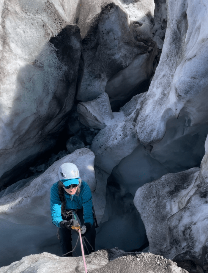 A woman abseils down a crevasse in a glacier wearing a helmet, sunglasses, a blue fleece, and black trousers. She is surrounded by grey and white ice.