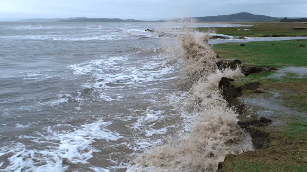 Small brown waves crash into the green coast, water splashes upwards as the they meet. The sky is grey and hills can be seen in the background.