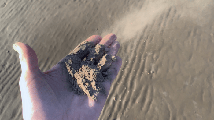 Over a sandy beach an open palm holds sand, grains fall through the fingers.