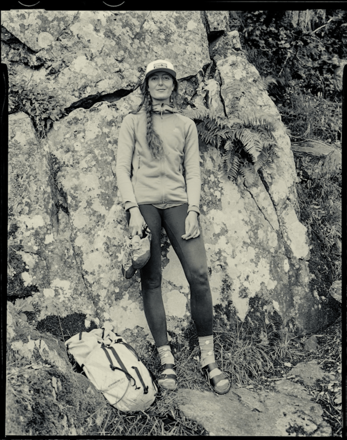A black and white image of a woman standing in front of a rock face, she wears outdoor gear, sandals, and is holding climbing shoes. There is a rucksack on the floor in front of her.