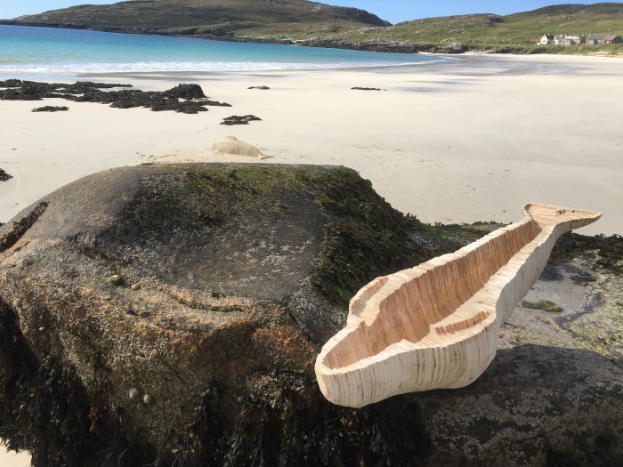 A sunny beach with white sand and a blue sea. The beach curves round the sea, with green hills in the background and some houses where the sand stops and the grass begins. In the foreground there is a large rock with a wooden mould of a whale lying on top of it.