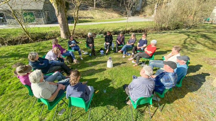 People sit in a circle having a discussion outdoors on green chairs, they are on grass and there is a stream and trees behind them. There is a slate building and small road in the background.