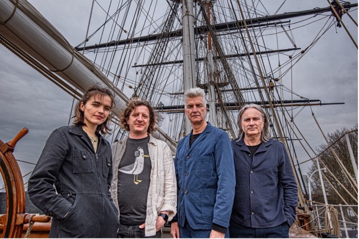 A white woman and three white men stand on the deck of a ship with the riggings behind them
