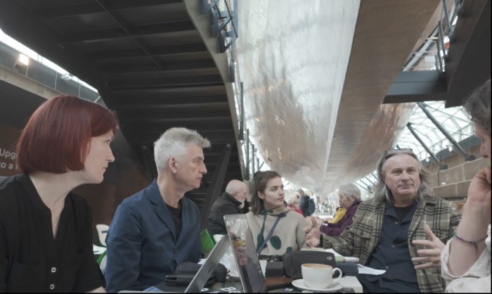 A group of 4 people talking around a table: suspended above them is the shiny hull of a large ship